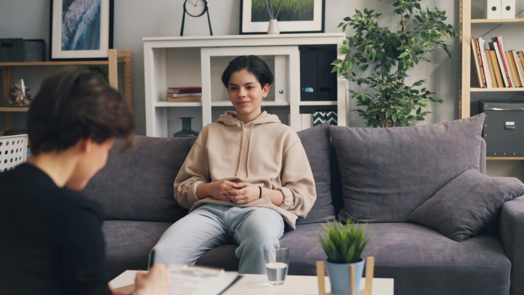 Teenager in therapy session sitting on a sofa, discussing with a professional therapist in a modern office setting.